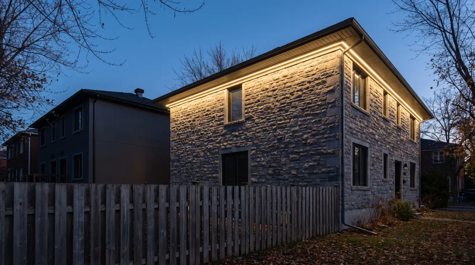 A two-storey grey stone house at dusk with a continuous warm-white line of light under the soffit washing the facade downward