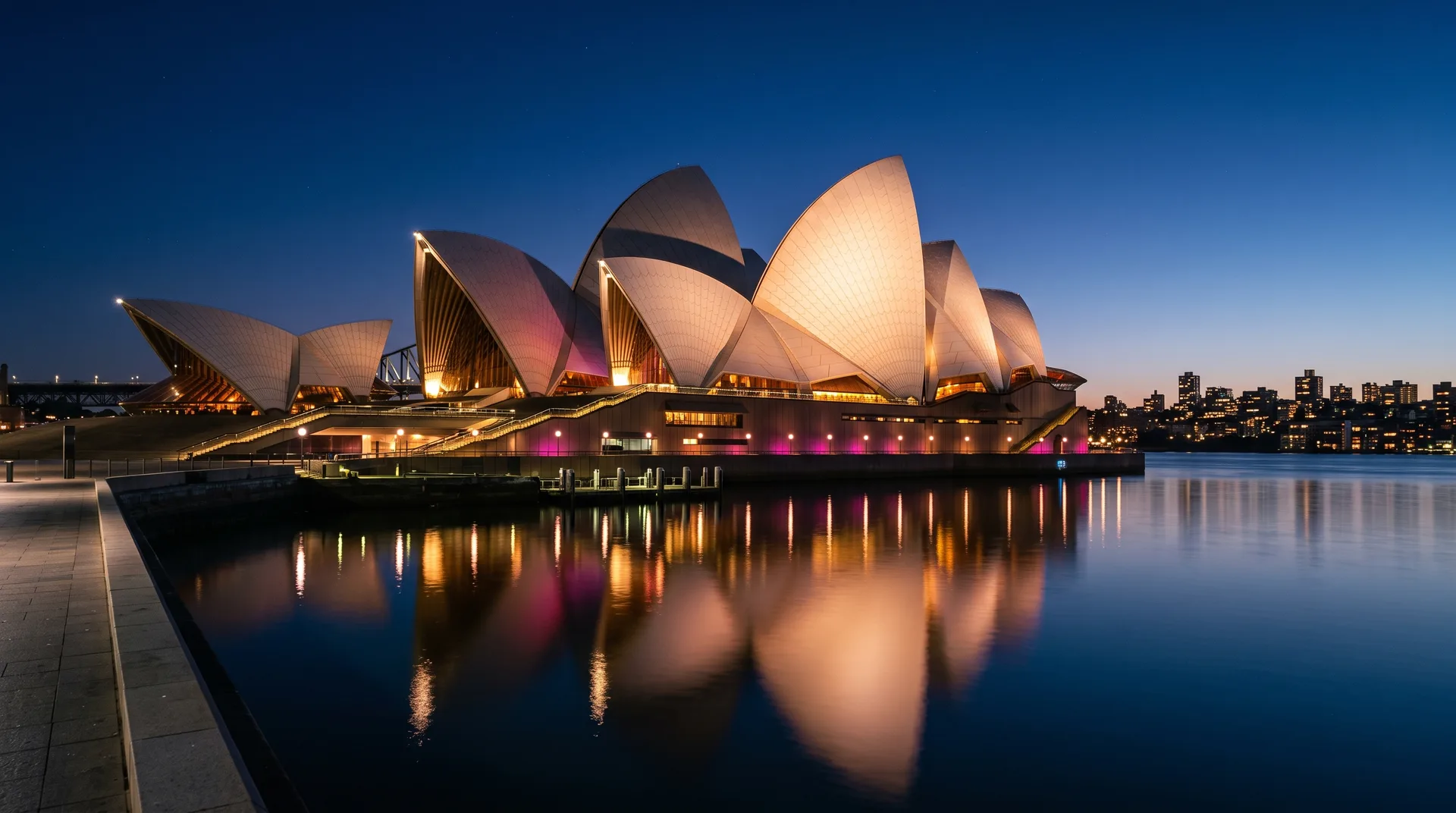 Night view of a large white shell-roofed building on the waterfront washed in coloured light