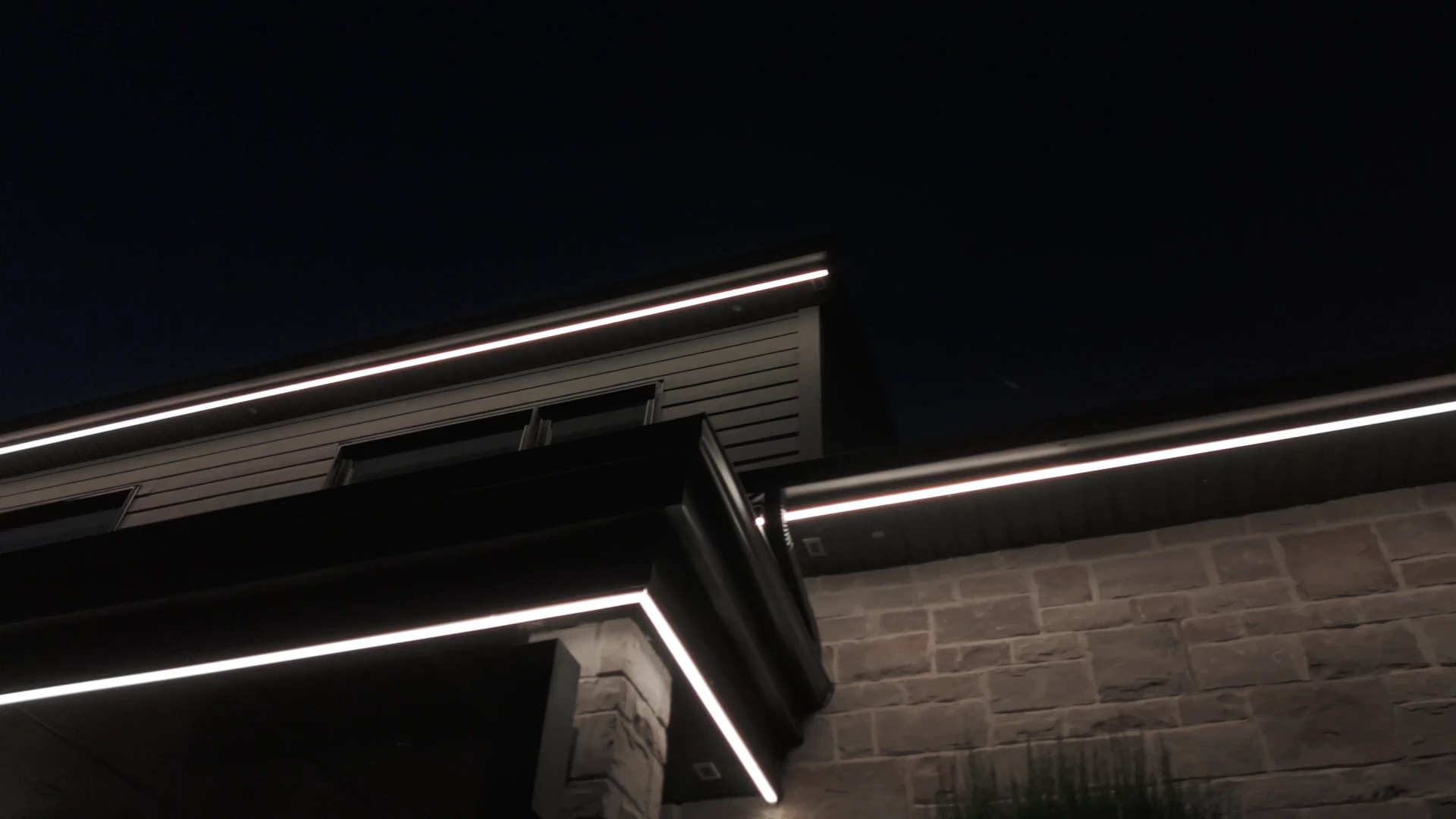 Night view up at a roofline: an unbroken warm-white LED line following the soffit, the overhang and the return above the porch, stone wall below