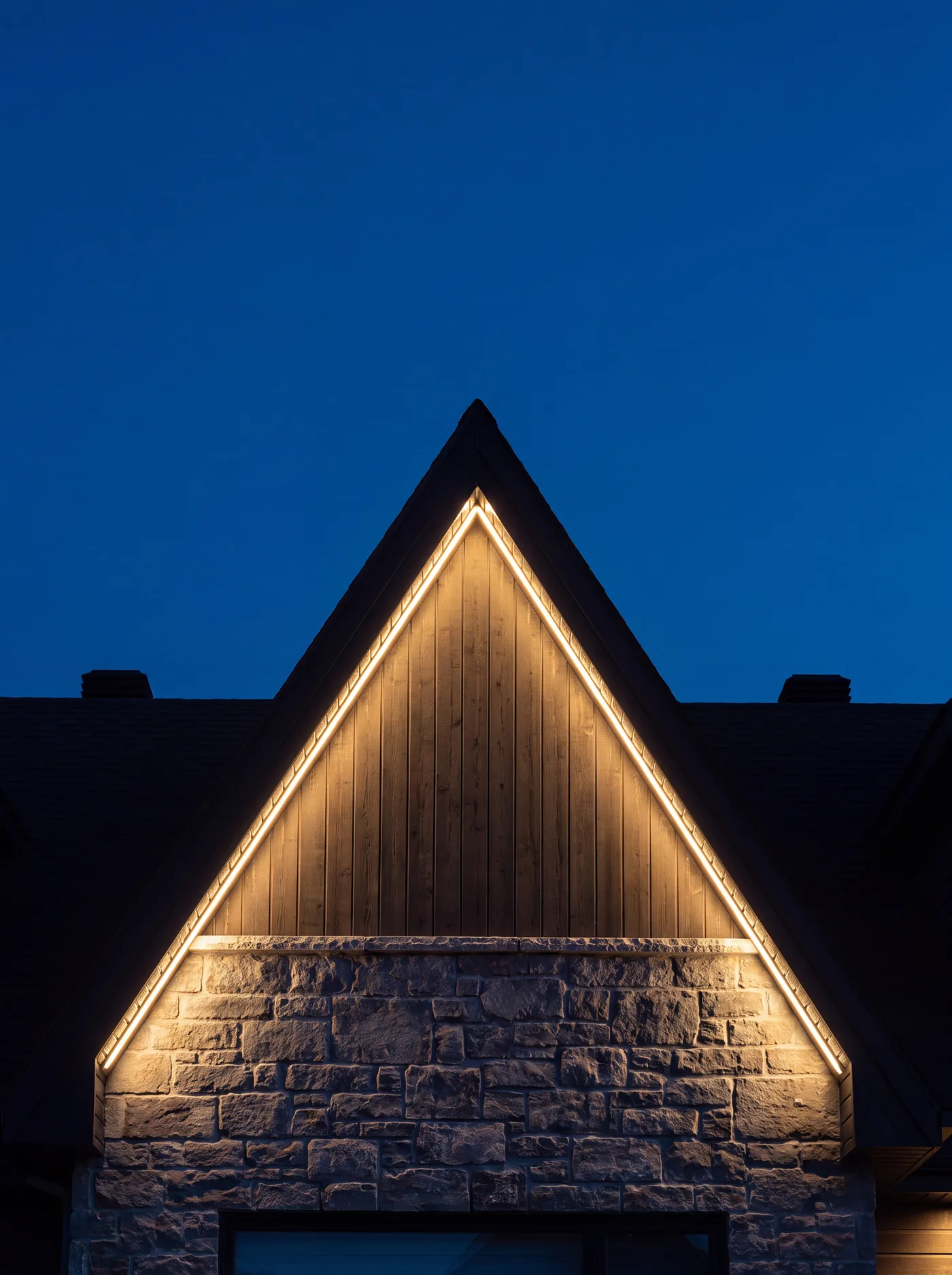 A house peak at night, the triangular top of a pitched roof outlined edge to edge in a continuous warm white LED line, wood siding above a stone base