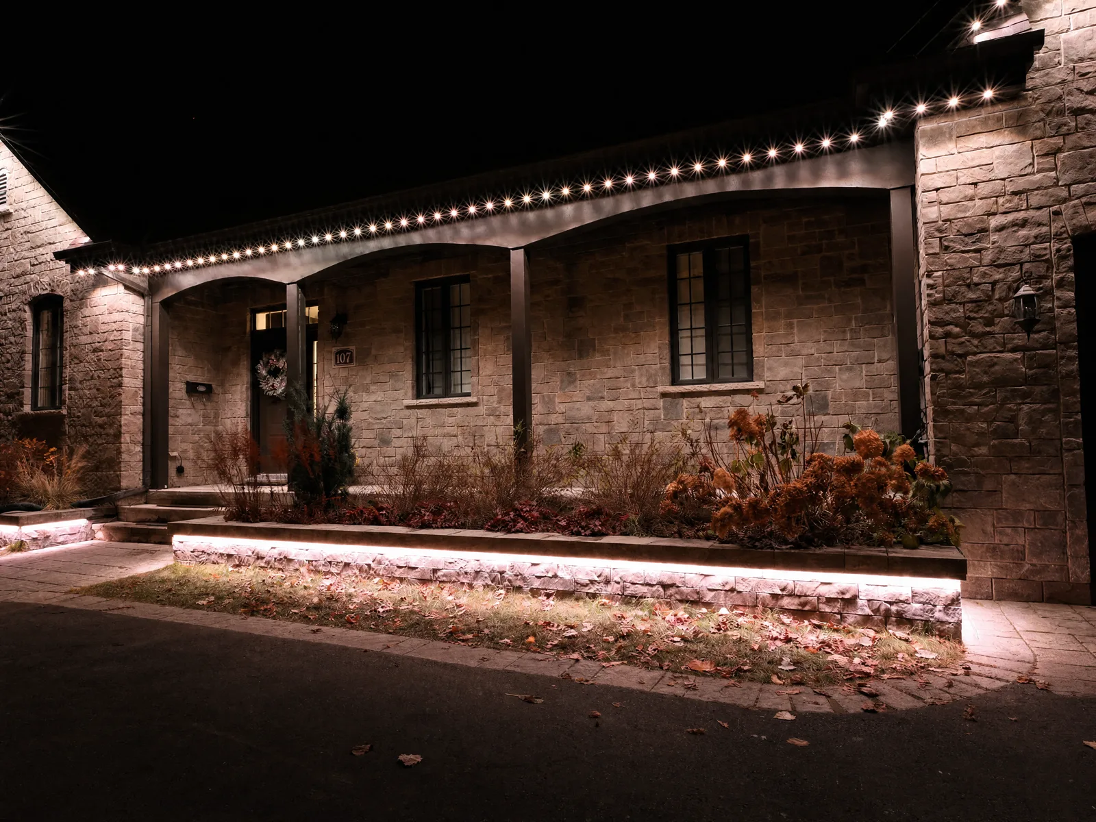 Stone home at night with warm-white permanent LED lighting tracing the porch arches and roofline, plus a warm wash along the garden wall — a Montréal-area install