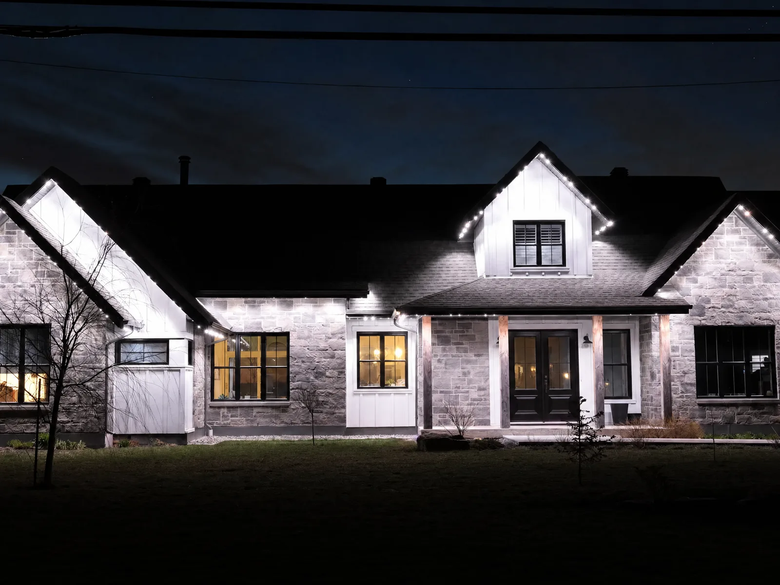 Grey-stone modern farmhouse at night with cool-white permanent LED lighting outlining every gable and roofline — a Québec home