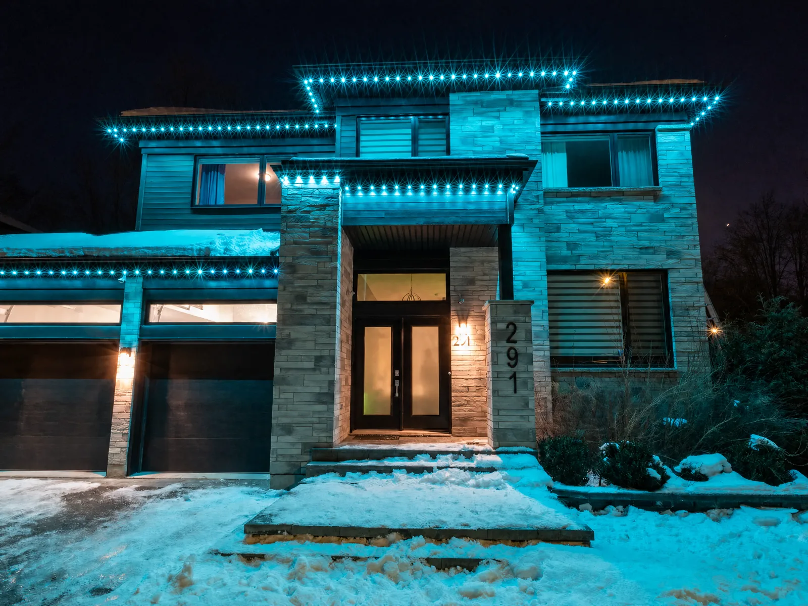 Modern stone home on a snowy winter night lit in vivid teal with permanent RGB LED tracing the rooflines — a Québec install