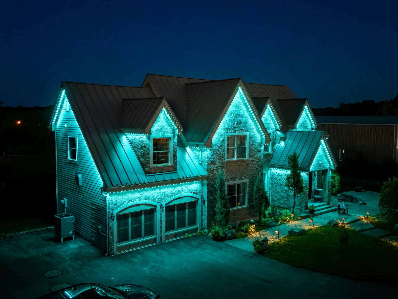 Large stone home with a metal roof at dusk, gables washed in teal-green permanent RGB LED with warm landscape path lighting — a Montréal-area property
